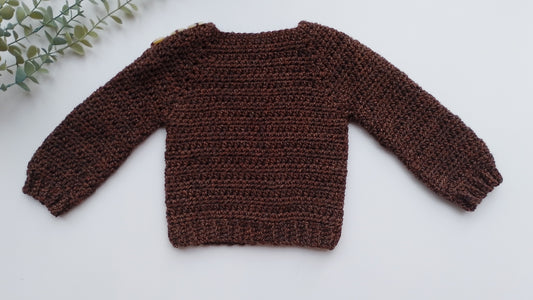 A dark brown crocheted long-sleeve sweater laid flat on a white background with a small green leafy sprig in the top-left corner.