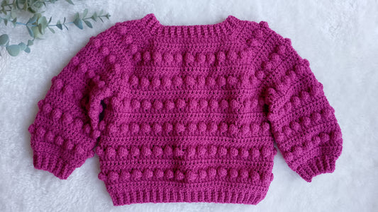 Showing the back view of a raspberry pink crocheted sweater with textured bobble stitches, laid flat on a white textured surface with a small green leafy sprig in the top right corner.