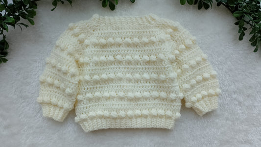 A cream-colored hand-crocheted baby sweater with textured bobble details, laid flat on a white faux fur surface with green leaves in the corners.