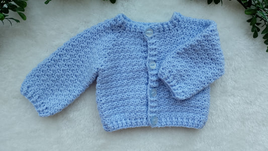 A light blue crocheted cardigan sweater with long sleeves and a button-up front, laid flat on a white textured surface with green leaves in the corners.