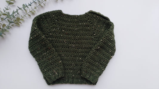 A dark green crocheted sweater with subtle gold flecks, laid flat on a white background with a decorative eucalyptus branch in the top-left corner.