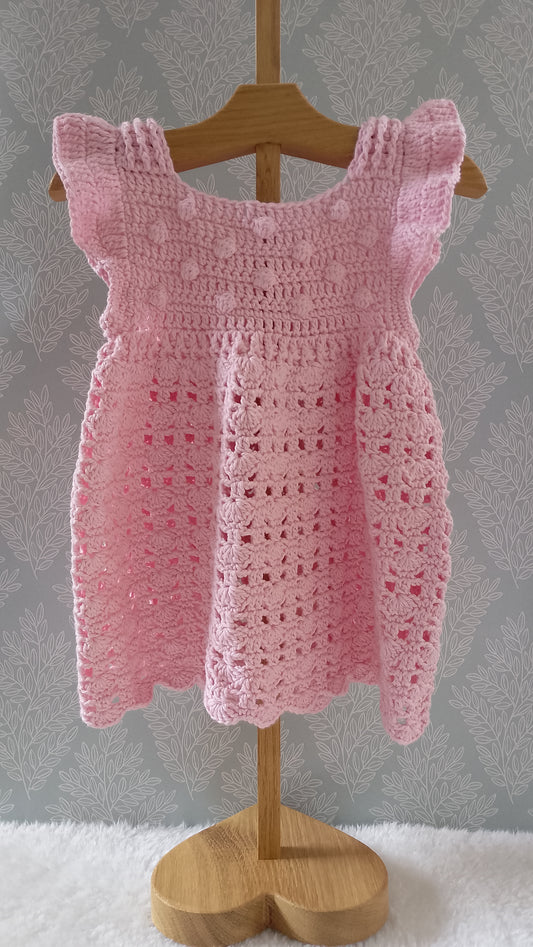 A pink crocheted baby dress with ruffled shoulder straps and a scalloped hem, displayed on a wooden hanger and stand against a gray patterned wall.