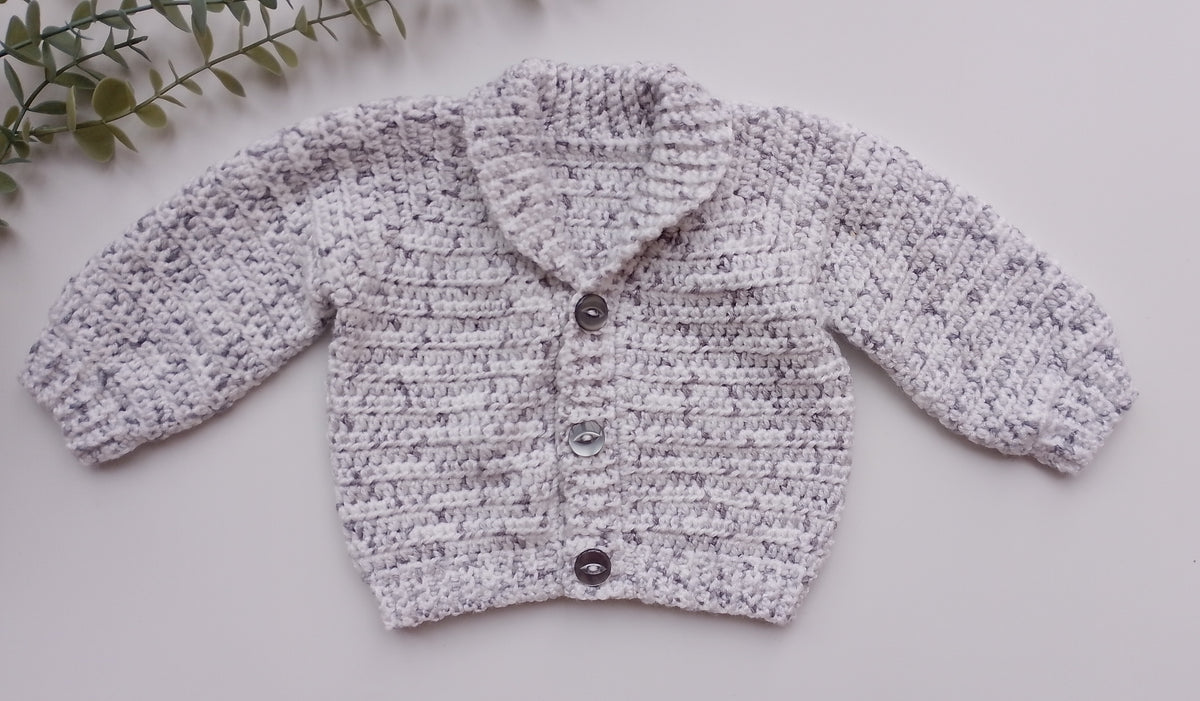 A light gray and white marled crochet cardigan sweater for a baby, featuring a pointed collar and three dark buttons, laid flat on a light gray background with a decorative green leaf sprig in the top-left corner.