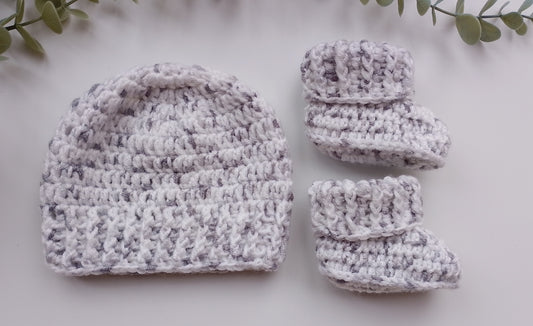 A handmade crochet baby hat and bootie set in a marled white and light gray yarn, styled flat-lay on a white surface with green leaves in the corners.