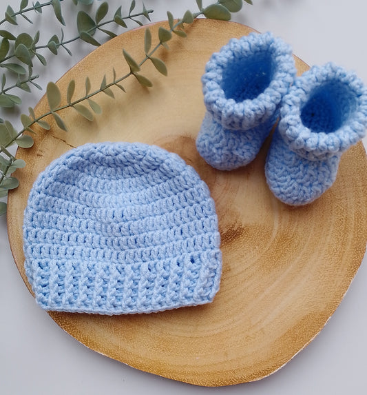 A light blue crocheted baby hat and matching booties arranged on a round wooden slice with green eucalyptus leaves, photographed on a light neutral background.