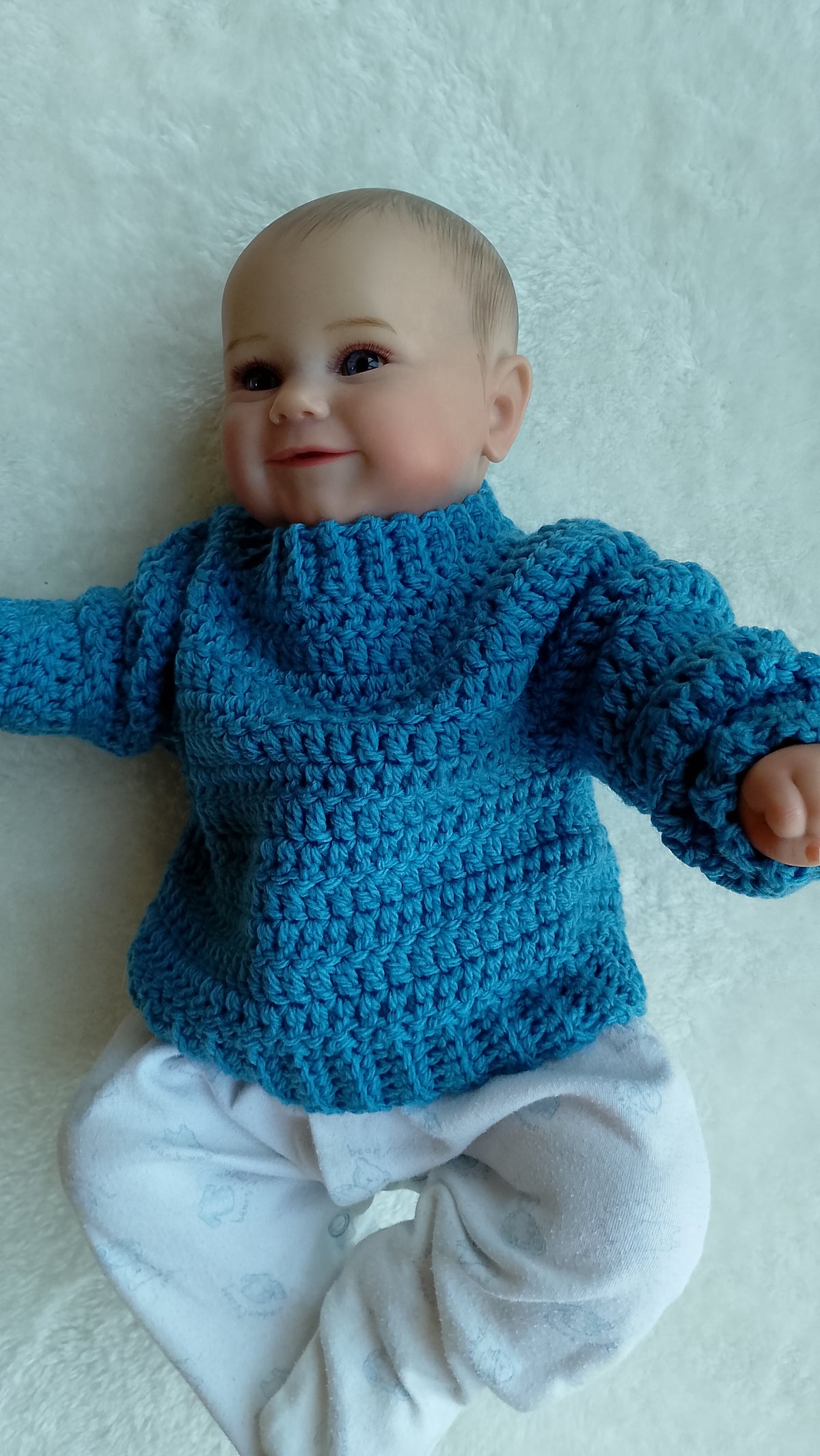 A close-up of a baby doll  lying on a soft white textured blanket, wearing a Cornish blue crocheted pullover jumper with a ribbed neckline.