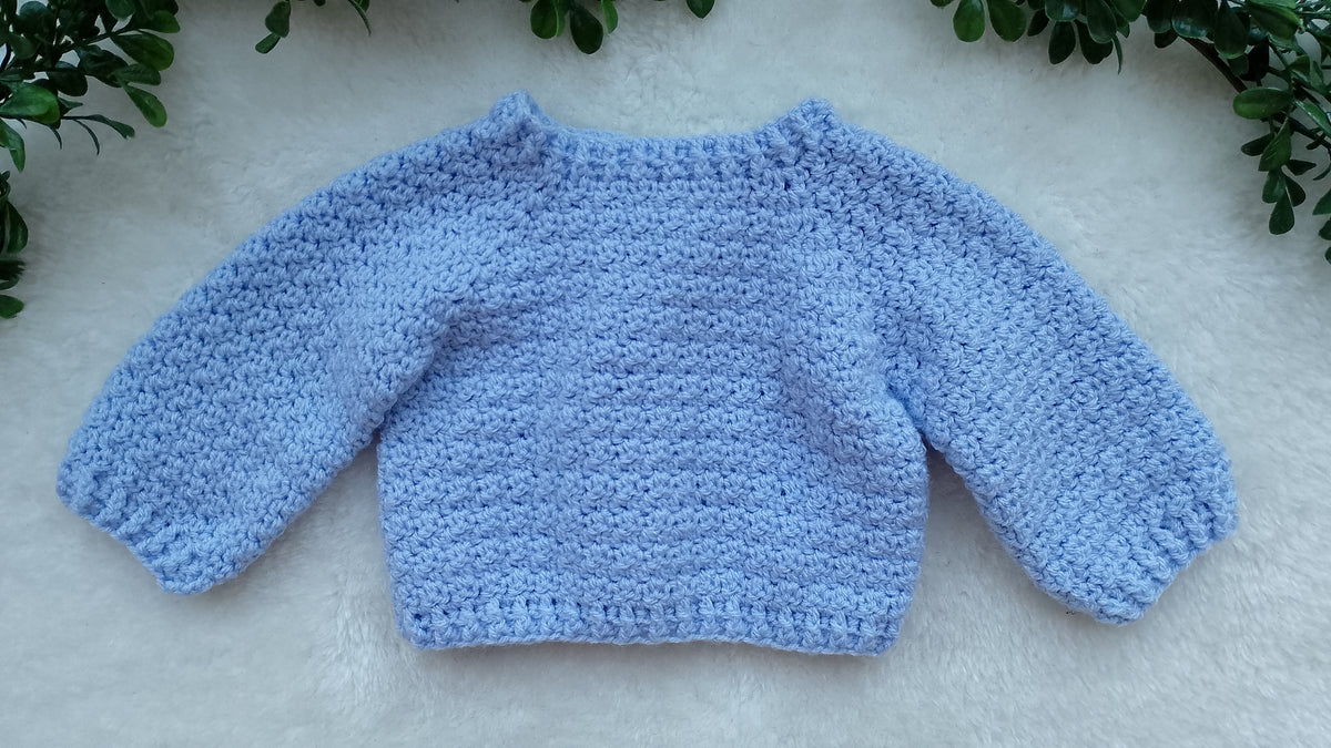A light blue crocheted baby sweater with long sleeves and a crew neck, laid flat on a white textured surface with green leaves in the corners.