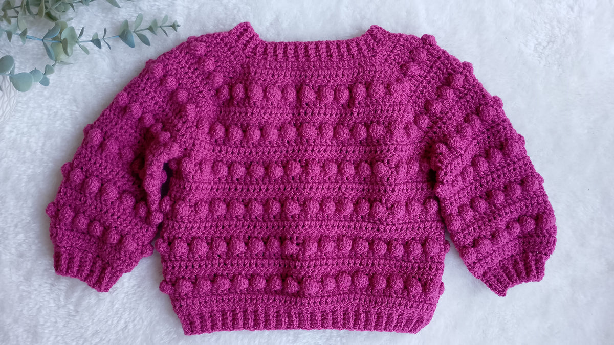 Showing the back view of a raspberry pink crocheted sweater with textured bobble stitches, laid flat on a white textured surface with a small green leafy sprig in the top right corner.