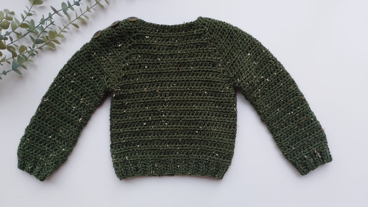 A dark green textured crochet sweater with subtle gold flecks, laid flat on a white background with a decorative green leafy branch in the top-left corner.