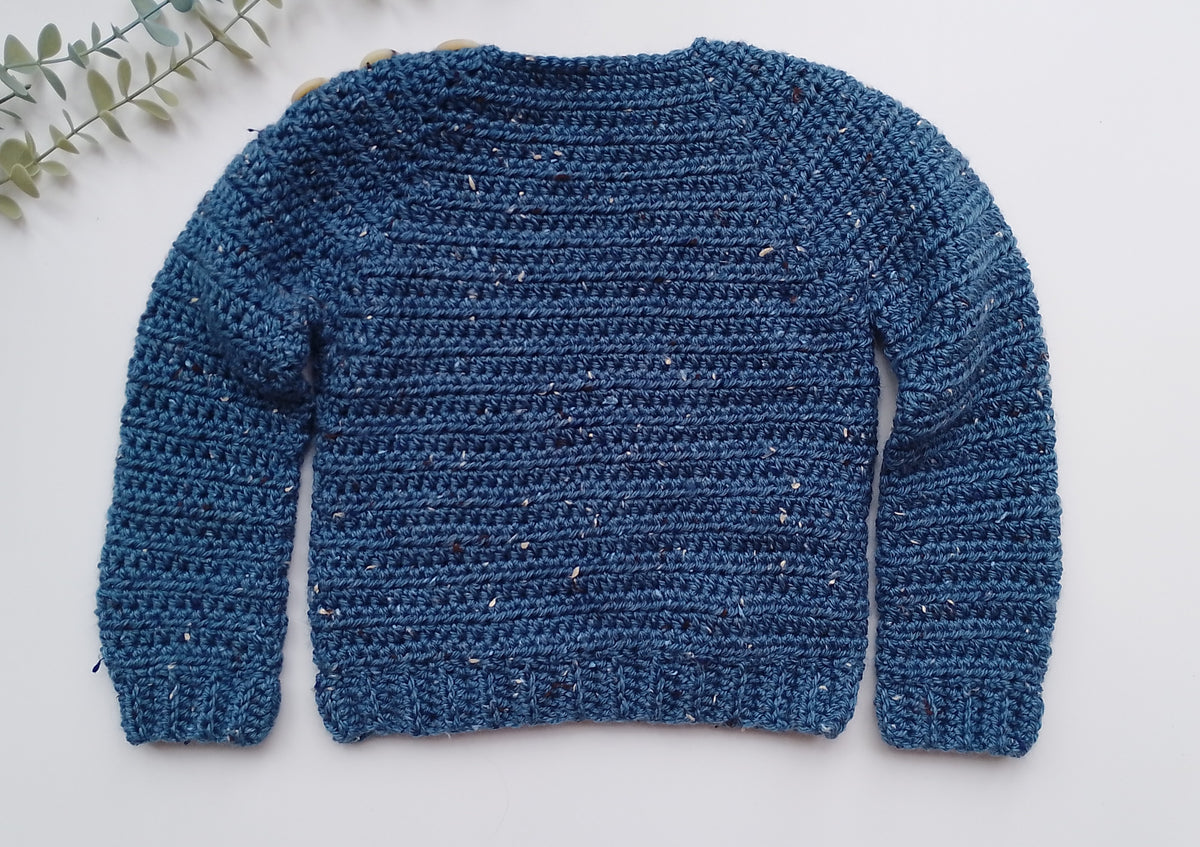A blue hand-crocheted knit sweater with long sleeves, laid flat on a white background with a small green leafy sprig in the top-left corner.