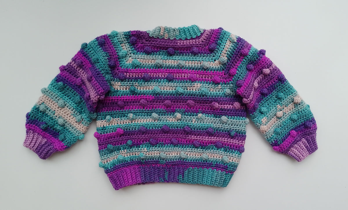 A handmade crocheted sweater with horizontal stripes of purple, teal,and beige, featuring  textured bobble design  details, laid flat on a light gray background.
