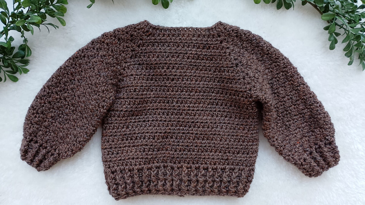 A brown chunky knit long-sleeve baby sweater laid flat on a white textured surface, framed by green leaves in the corners.
