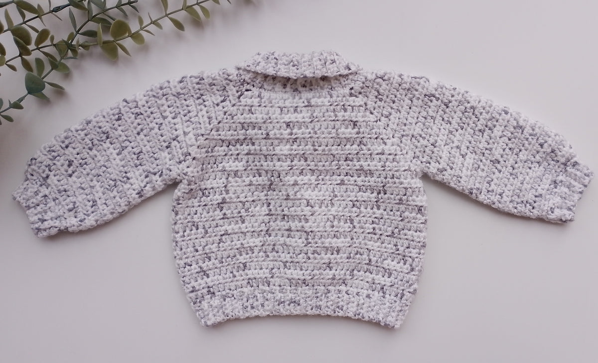 A light gray and white marled knit sweater with long sleeves, laid flat on a light gray surface with a green leafy sprig in the top-left corner.