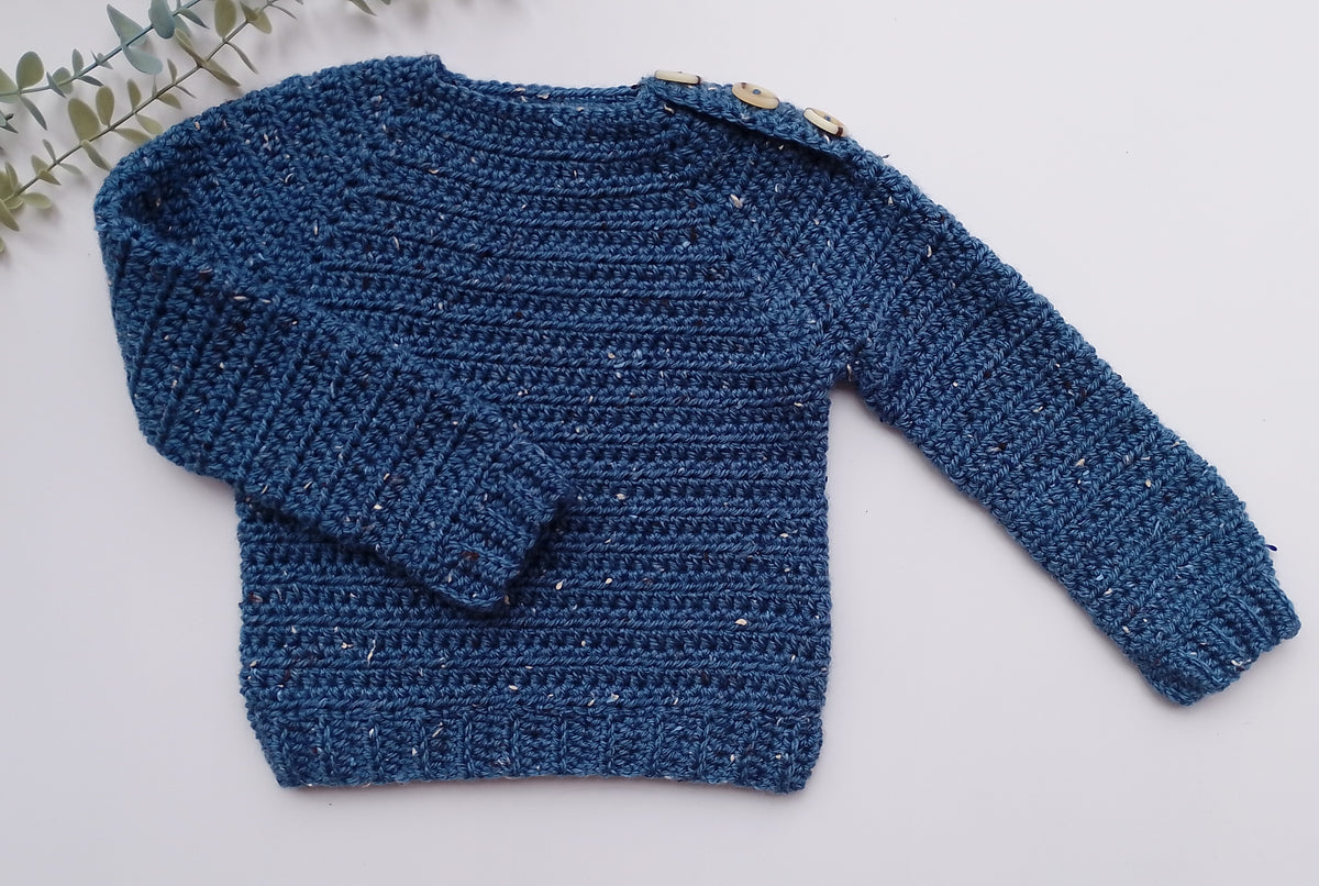 A textured blue crochet sweater with long sleeves and decorative wooden buttons on the shoulder, laid flat on a light gray background with a small green leafy sprig in the top-left corner.