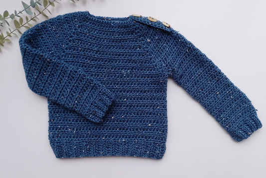 A textured blue crochet sweater with long sleeves and decorative wooden buttons on the shoulder, laid flat on a light gray background with a small green leafy sprig in the top-left corner.