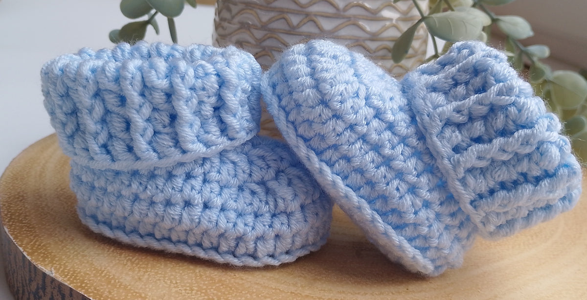 A pair of light blue crocheted baby booties displayed on a round wooden slice with a neutral patterned vase and green leaves in the background.