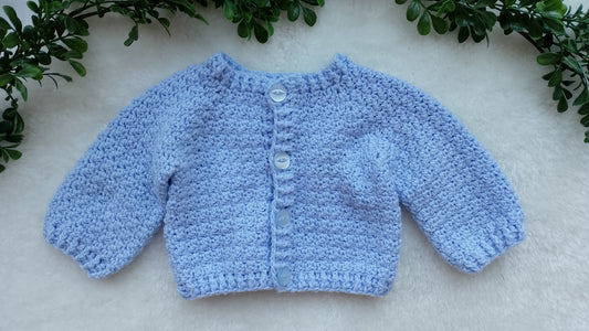 A light blue crocheted cardigan sweater for a baby or toddler, featuring four front buttons, laid flat on a white textured surface with green foliage framing the top corners.
