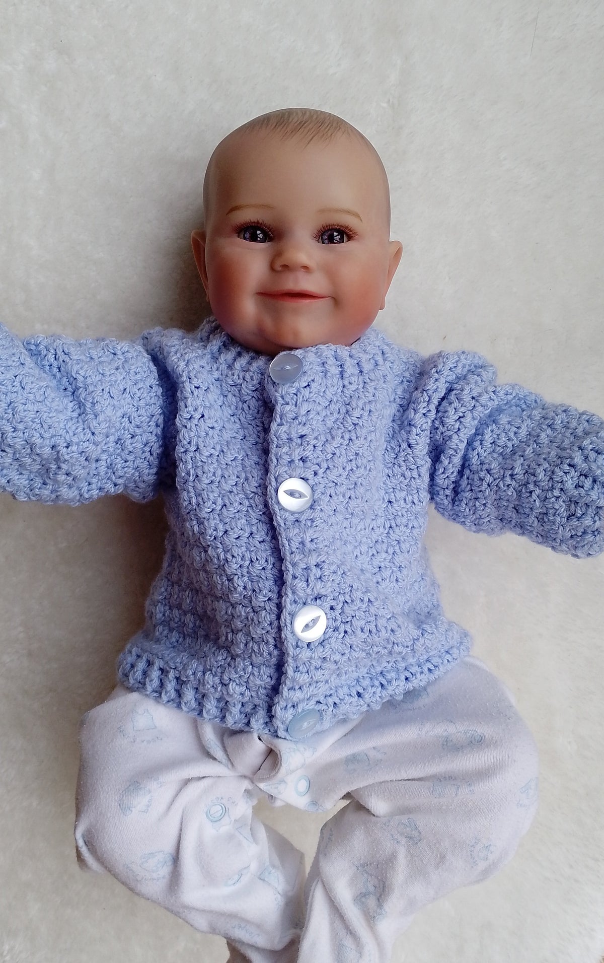 A baby doll with realistic skin and hair wearing a light blue crochet cardigan over a white onesie, posed against a soft beige fabric background.