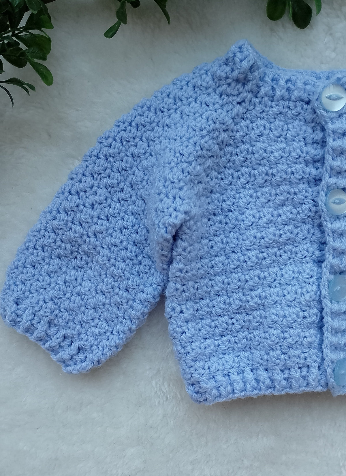 A light blue crocheted cardigan or sweater laid flat showing the sleeve texture on a white faux-fur surface, with green leaves in the top corners.