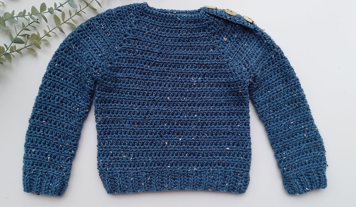 A textured blue crocheted knit sweater with long sleeves and a crew neck, laid flat on a white background with a decorative green leafy sprig in the top-left corner.