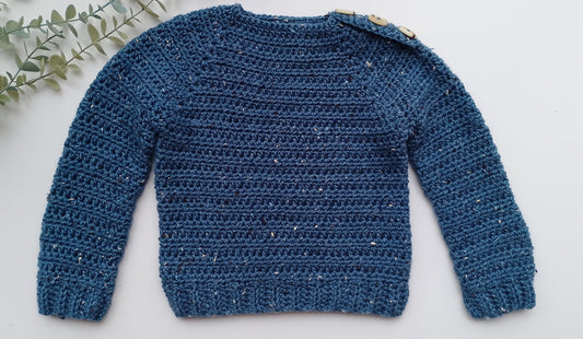 A textured blue crocheted knit sweater with long sleeves and a crew neck, laid flat on a white background with a decorative green leafy sprig in the top-left corner.
