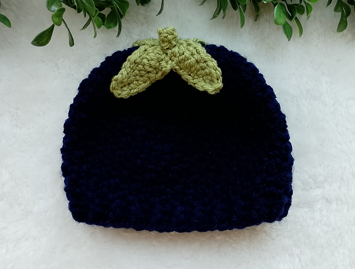 A dark navy blue crocheted beanie hat with a light olive green leaf-shaped accent on top, displayed on a white textured surface with green leaves at the top edge.