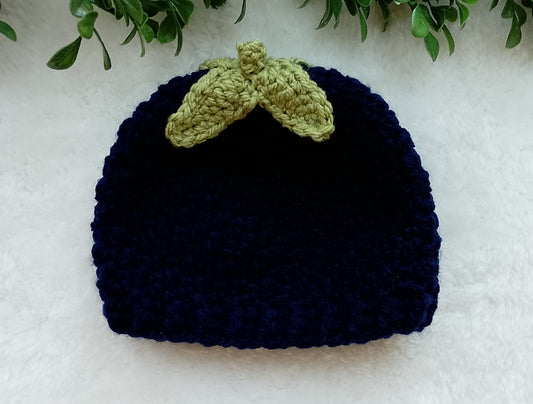 A dark navy blue crocheted beanie hat with a light olive green leaf-shaped accent on top, displayed on a white textured surface with green leaves at the top edge.
