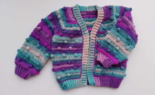 A handmade, hand-crocheted cardigan sweater featuring a colorful striped pattern in shades of purple, teal, with pom-pom accents, laid flat on a light gray background.