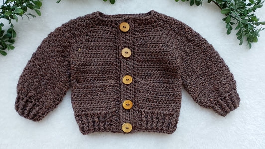 A brown crocheted cardigan sweater with four wooden buttons, laid flat on a white textured surface with green leaves in the corners.