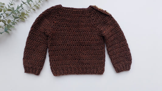 A dark brown textured crochet sweater for toddlers or babies, laid flat on a white background with a decorative green eucalyptus branch in the top-left corner.