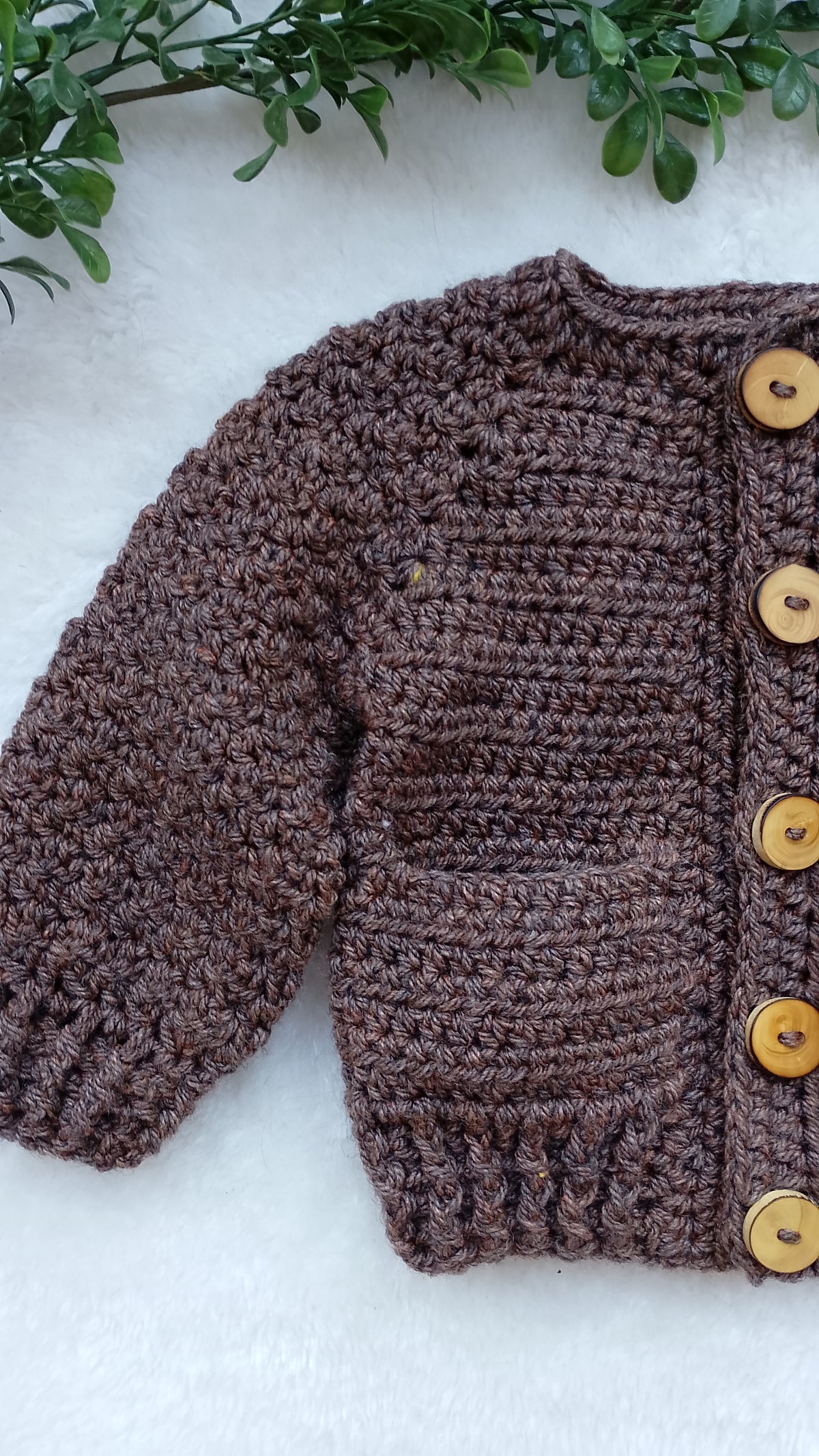 A brown chunky knit cardigan sweater with wooden buttons, laid flat on a white textured surface with green leaves at the top.