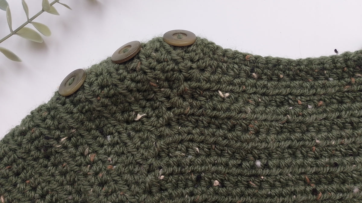 Close-up of a textured olive green crocheted sweater or pullover featuring three green buttons along the neckline, set against a clean white background with a small leafy sprig in the corner.