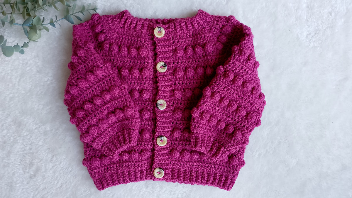 A raspberry pink crocheted cardigan sweater with textured bobble details, laid flat on a white textured surface with decorative green leaves in the top-left corner.