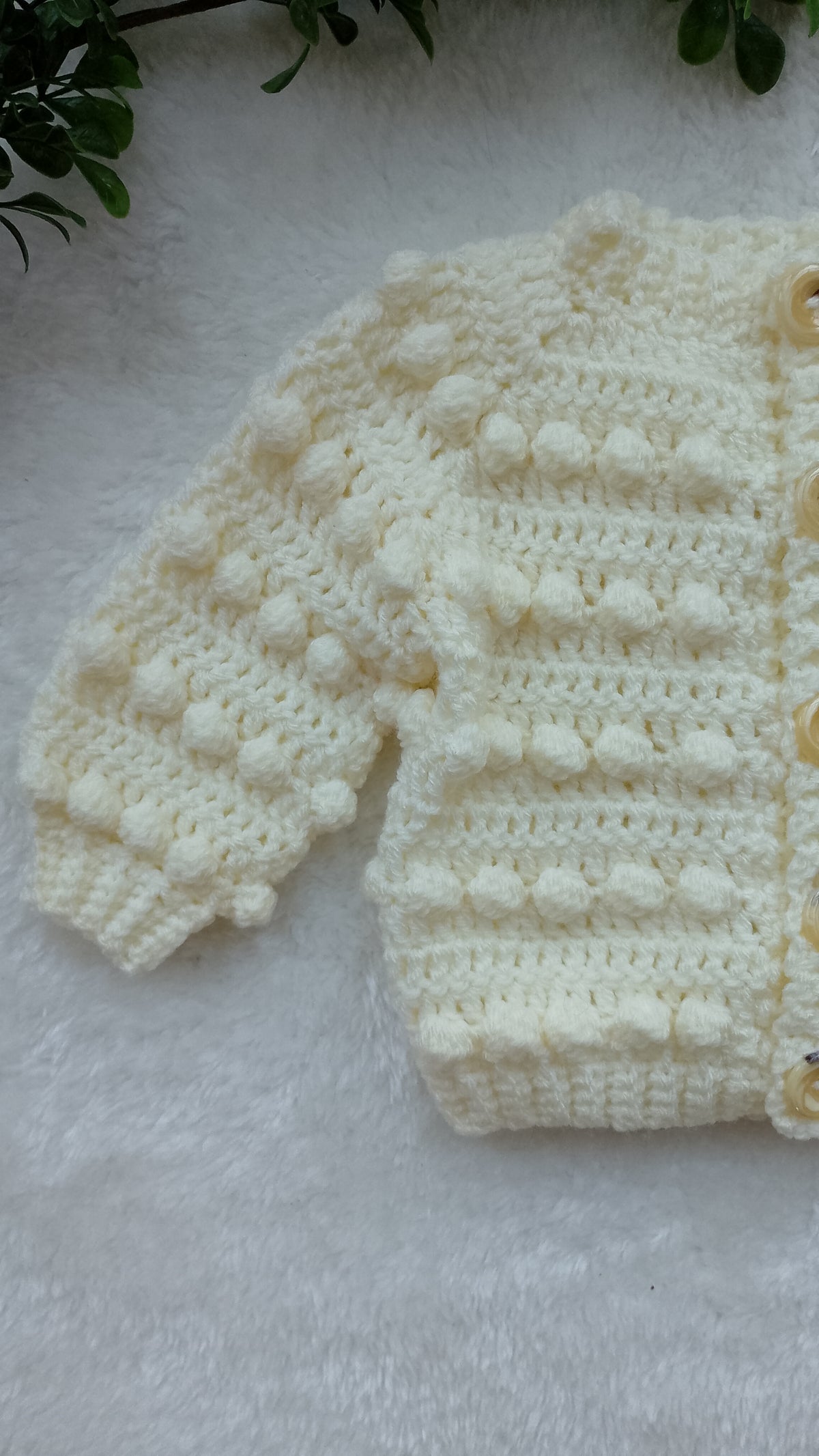 A cream-colored crocheted baby cardigan with textured bobble stitches, laid flat on a white faux fur surface with green leaves in the top corner.