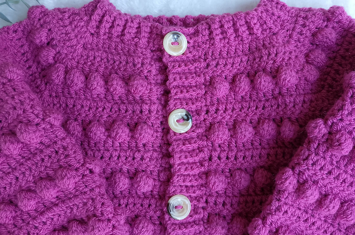 A close-up of a crochet cardigan in a raspberry pink, featuring a textured bobble pattern and decorative cream-colored buttons.