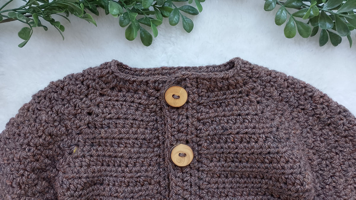 A close-up of a brown chunky crochet cardigan  laid flat on a white textured surface, with two round wooden buttons at the front and green leaves along the top edge.