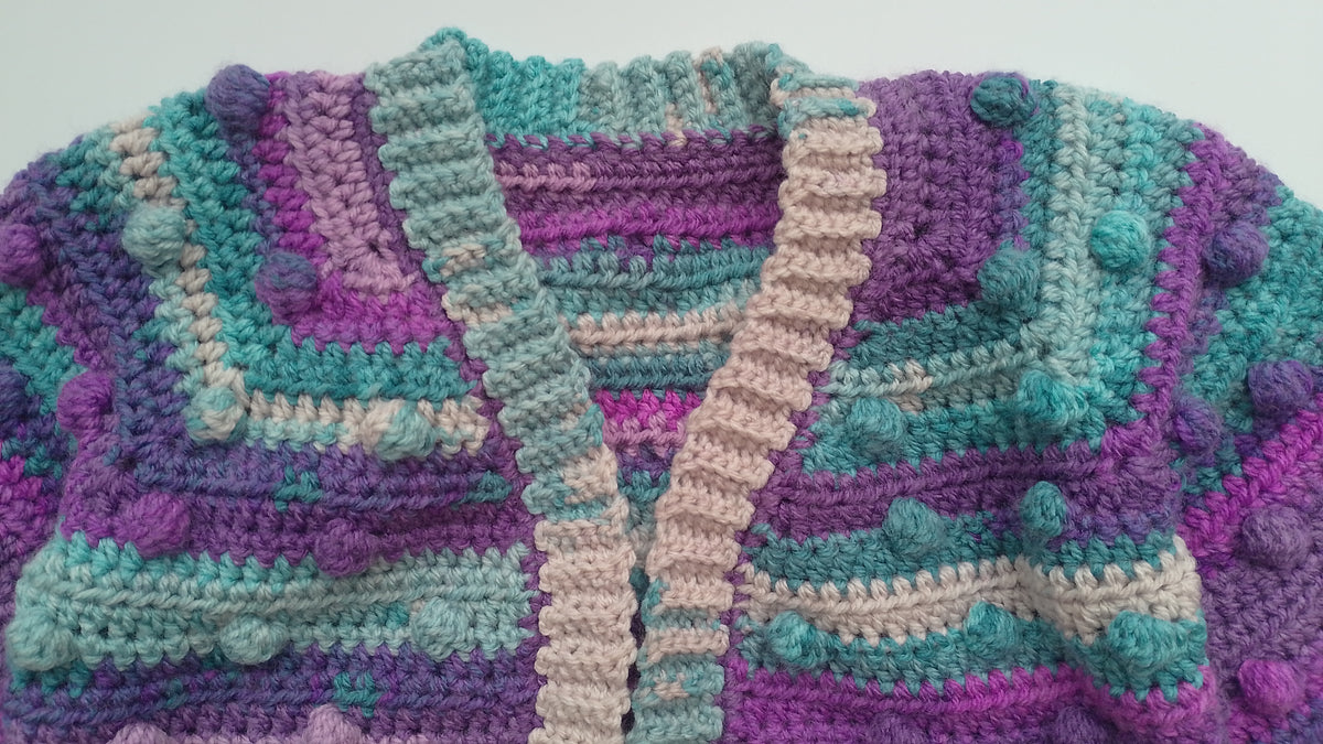 A close-up of a hand-crocheted cardigan featuring textured stripes in purple, teal and beige, accented with small bobbles throughout.