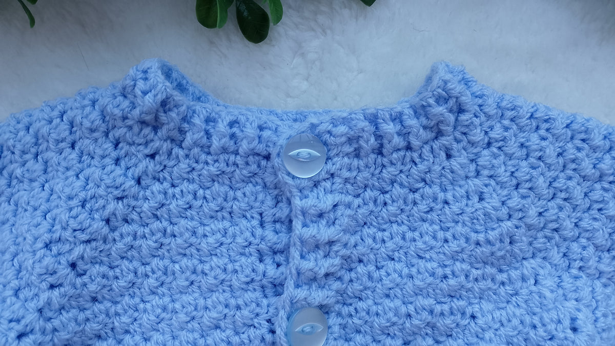 A close-up of a light blue crocheted cardigan or sweater with a textured pattern and  light blue buttons, laid flat on a white textured surface with a small green leaf at the top edge.
