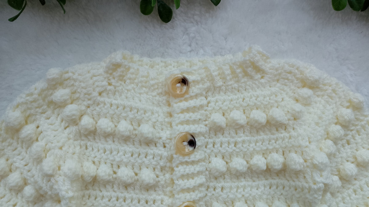 A close-up of a cream-colored, hand-crocheted cardigan featuring textured bumpy crochet patterns and vintage buttons, laid flat on a white textured surface.