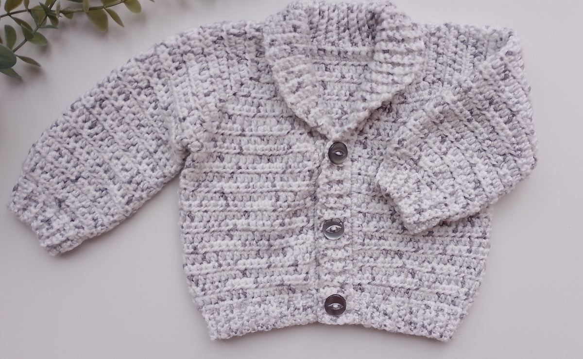 A light gray and white marled crochet cardigan sweater with three dark buttons, laid flat on a light gray surface with a small green leafy sprig in the top-left corner.