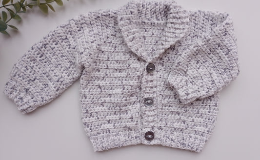 A light gray and white marled crochet cardigan sweater with three dark buttons, laid flat on a light gray surface with a small green leafy sprig in the top-left corner.