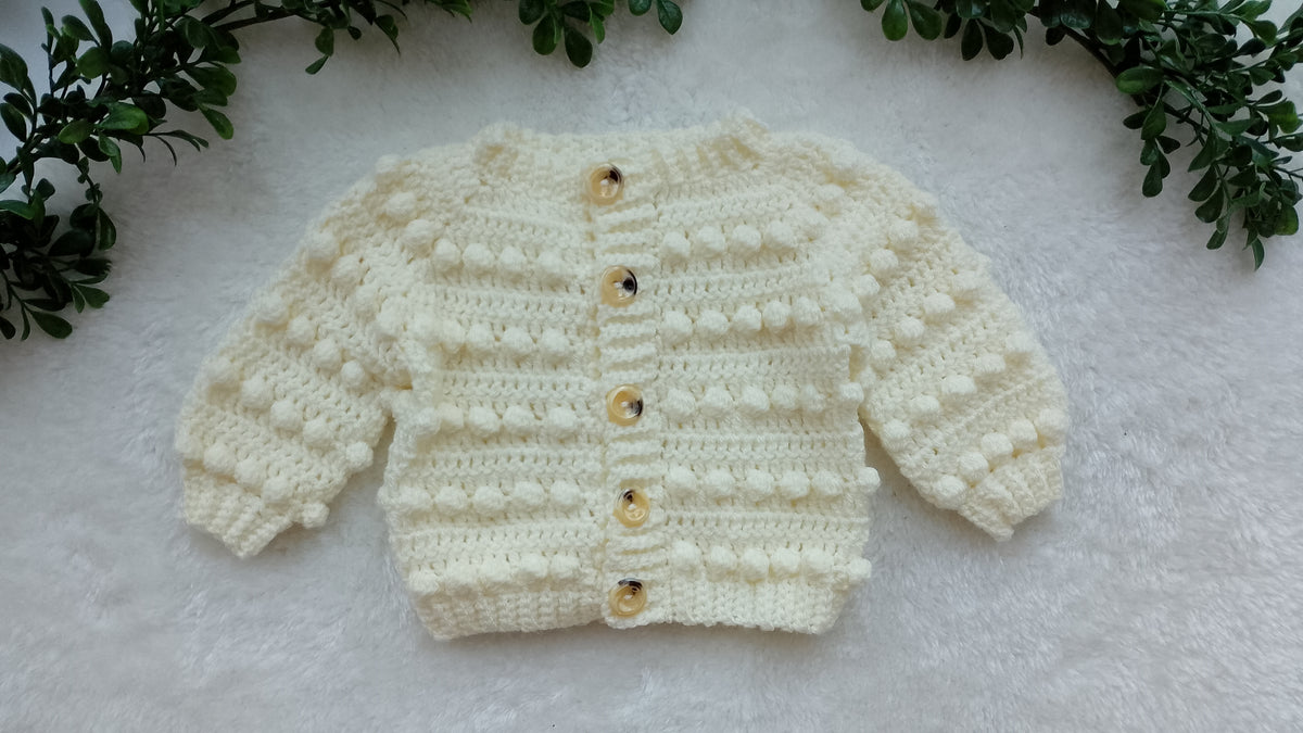 A cream-colored hand-crocheted baby sweater with textured bobble details and vintage style buttons, laid flat on a white faux fur surface with green leaves in the corners.