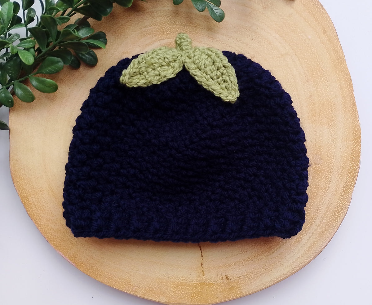 A dark navy blue crocheted beanie hat in a blueberry hat style with two light green leaf accents on top, displayed on a round wooden slice with green leaves in the corner.