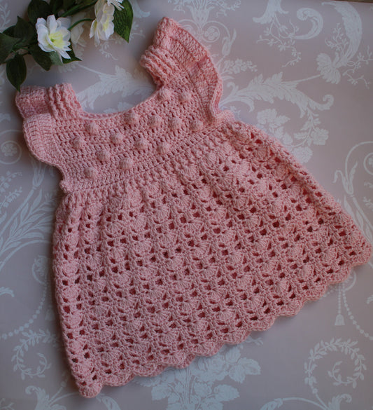 A handmade, light pink crocheted baby dress with short sleeves and a scalloped hem, laid flat on a beige damask-patterned surface with white roses in the corner.