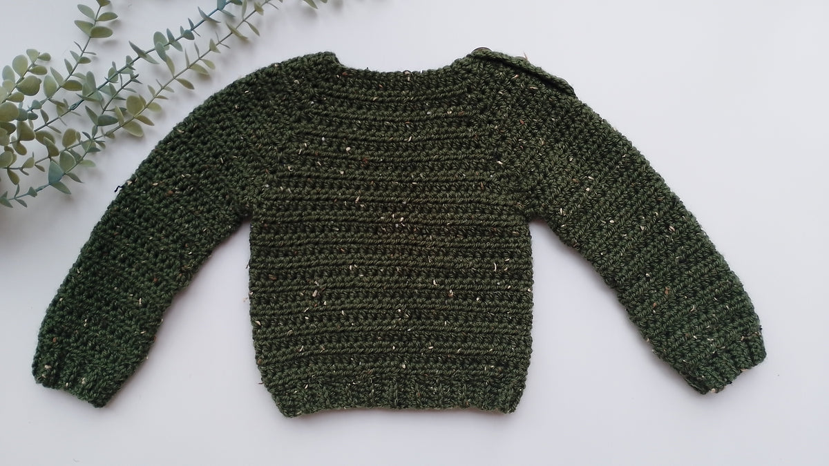 An olive green crocheted long-sleeve sweater with subtle gold/bronze flecks, laid flat on a white background with a decorative eucalyptus sprig in the top-left corner.