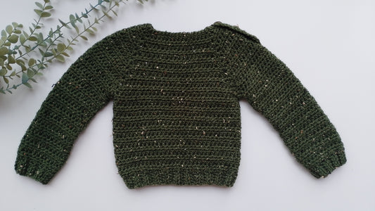 An olive green crocheted long-sleeve sweater with subtle gold/bronze flecks, laid flat on a white background with a decorative eucalyptus sprig in the top-left corner.