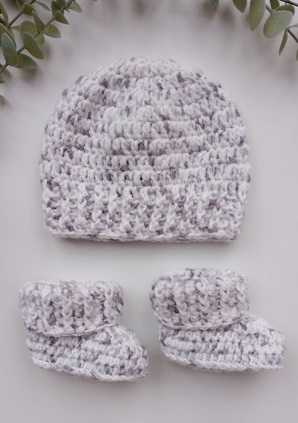 A handmade crochet baby hat and matching booties in a marled white and light gray yarn, arranged on a light gray background with green leaves in the corners.