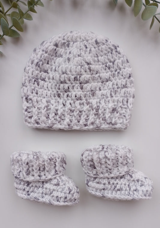A handmade crochet baby hat and matching booties in a marled white and light gray yarn, arranged on a light gray background with green leaves in the corners.
