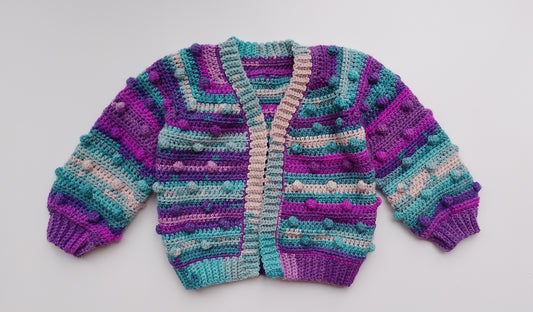 A handmade, hand-crocheted cardigan with long sleeves, featuring horizontal stripes in purple, teal, and beige, accented with raised pom-pom details. It is displayed flat against a light gray background.