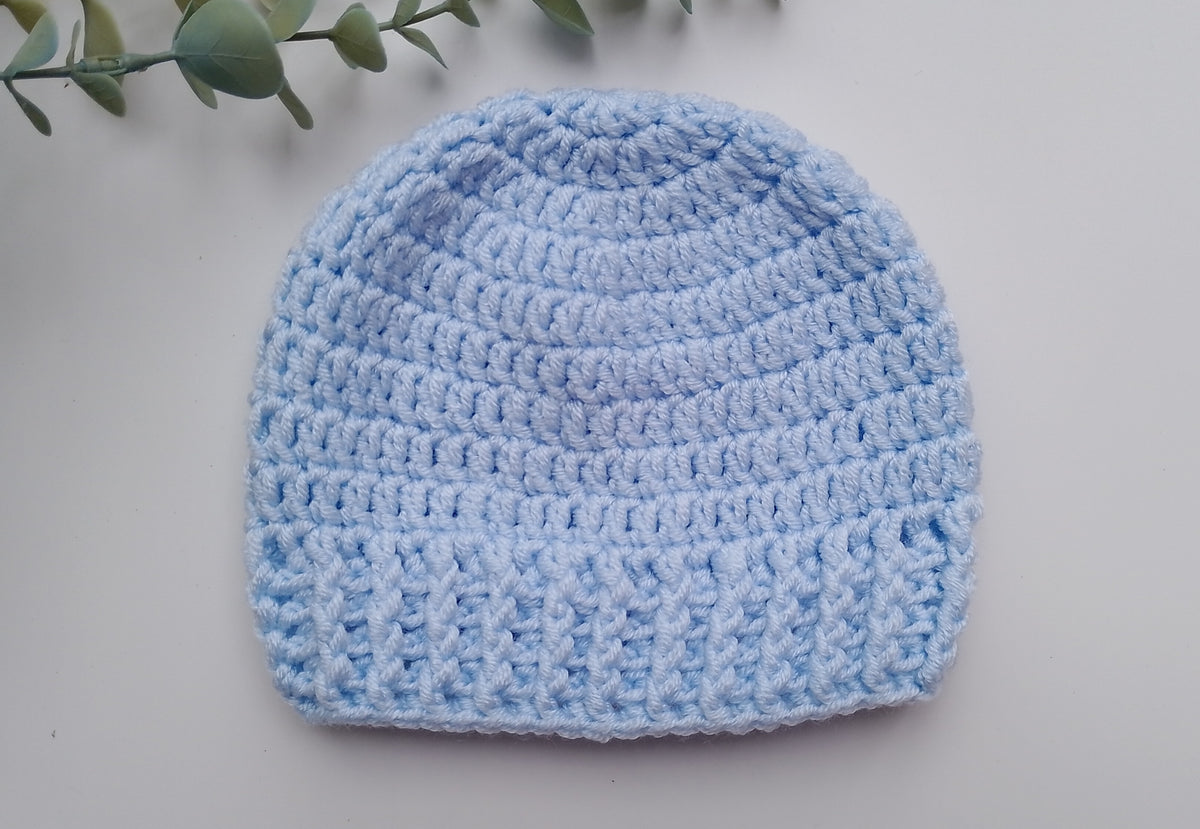 A light blue crocheted beanie hat laid flat on a light gray surface, with a small green leafy branch visible in the top-left corner.